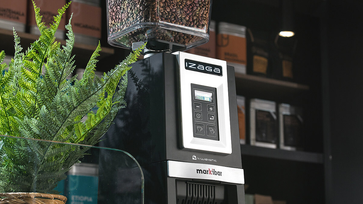 Markibar Izaga espresso grinder with a clear bean hopper on a café counter next to a potted plant, with coffee bags displayed in the background.