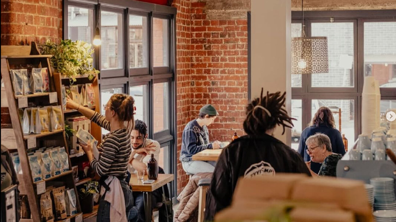 Cozy brick-walled coffee shop interior with customers seated at tables and a barista organizing coffee bags on a display shelf.