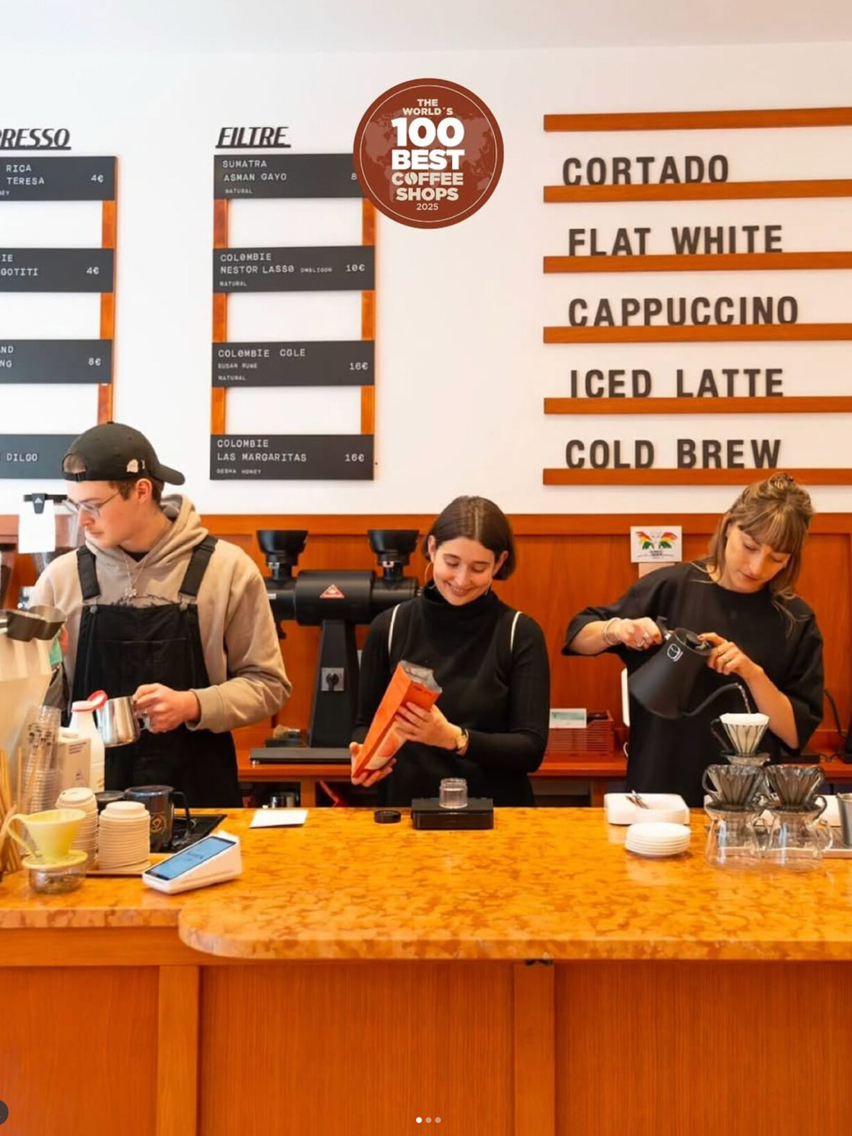 Three baristas prepare drinks behind a coffee bar with a menu board listing espresso and filter options and classic café drinks.