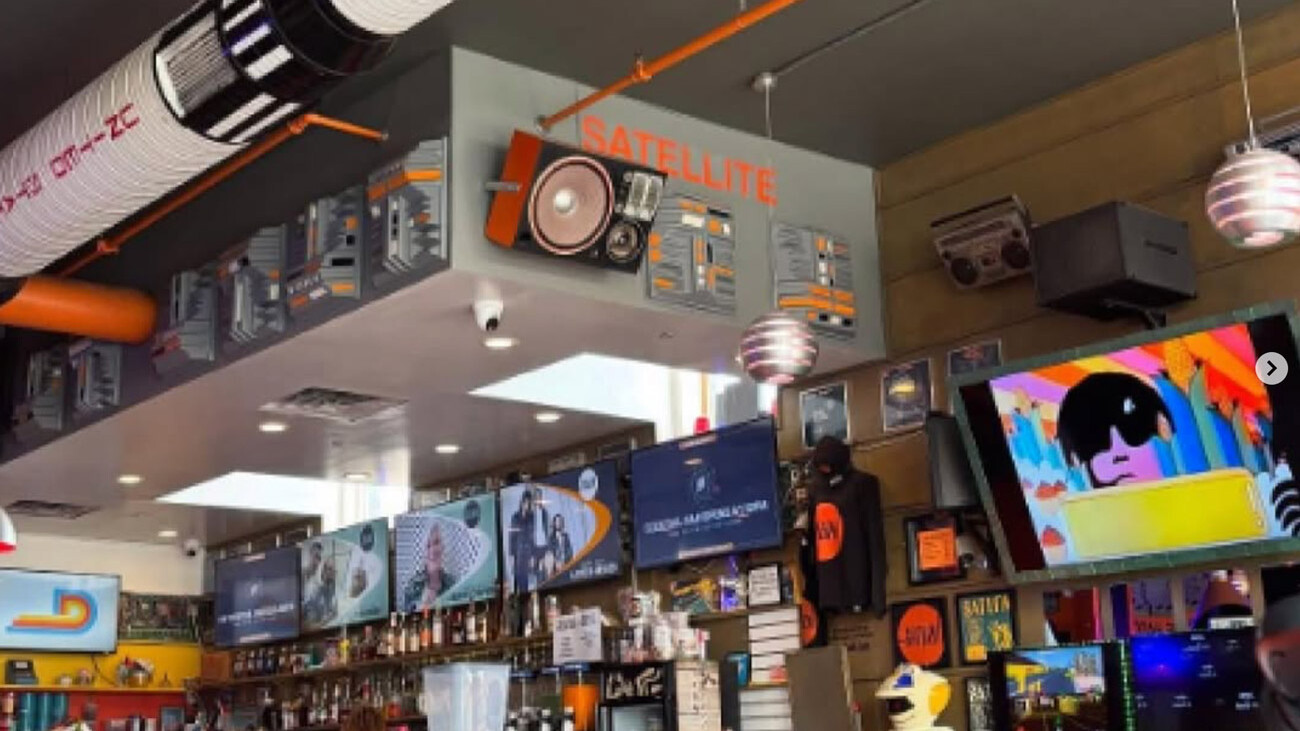 Bright, retro-style coffee shop interior with orange stools and tables, multiple screens behind the counter, and a “Satellite” sign hanging from the ceiling.