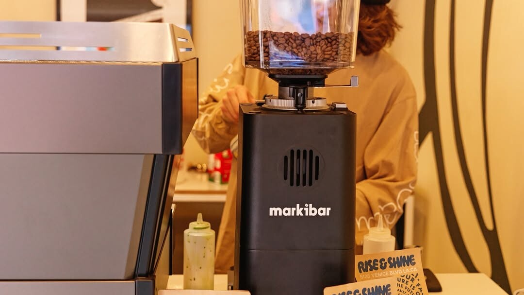 Markibar espresso grinder with coffee beans in the hopper next to an espresso machine, coffee bags, and cappuccino cups on a café counter.