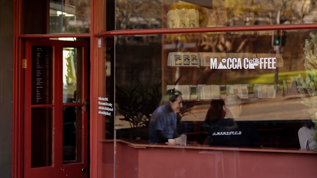 Exterior of a coffee shop with red-framed windows, hanging signage, and customers seated inside by the front window.
