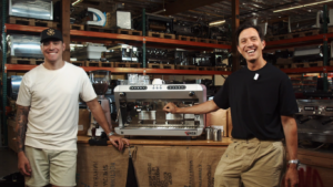Two men smiling in a coffee equipment warehouse, standing beside a Wega commercial espresso machine and grinder setup on a burlap-covered counter.