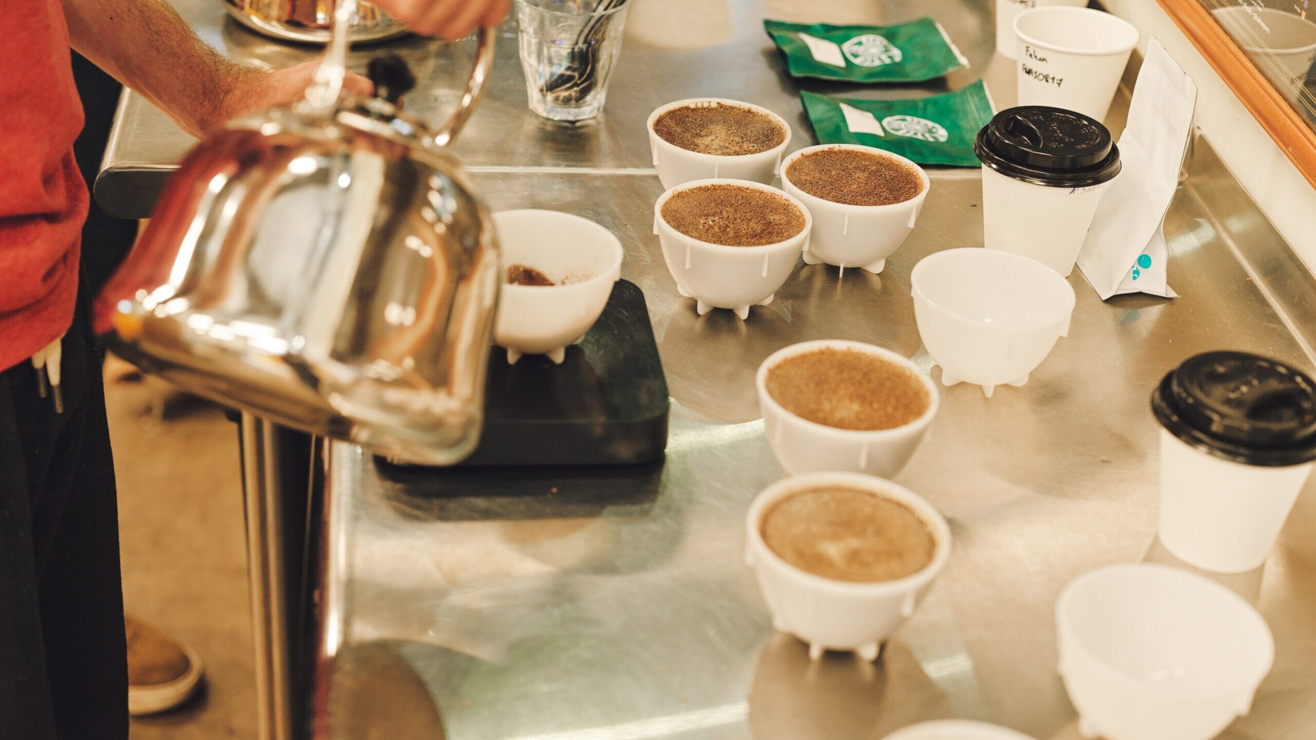 Barista pours hot water from a kettle into coffee cupping bowls lined up on a stainless steel table during a tasting session.