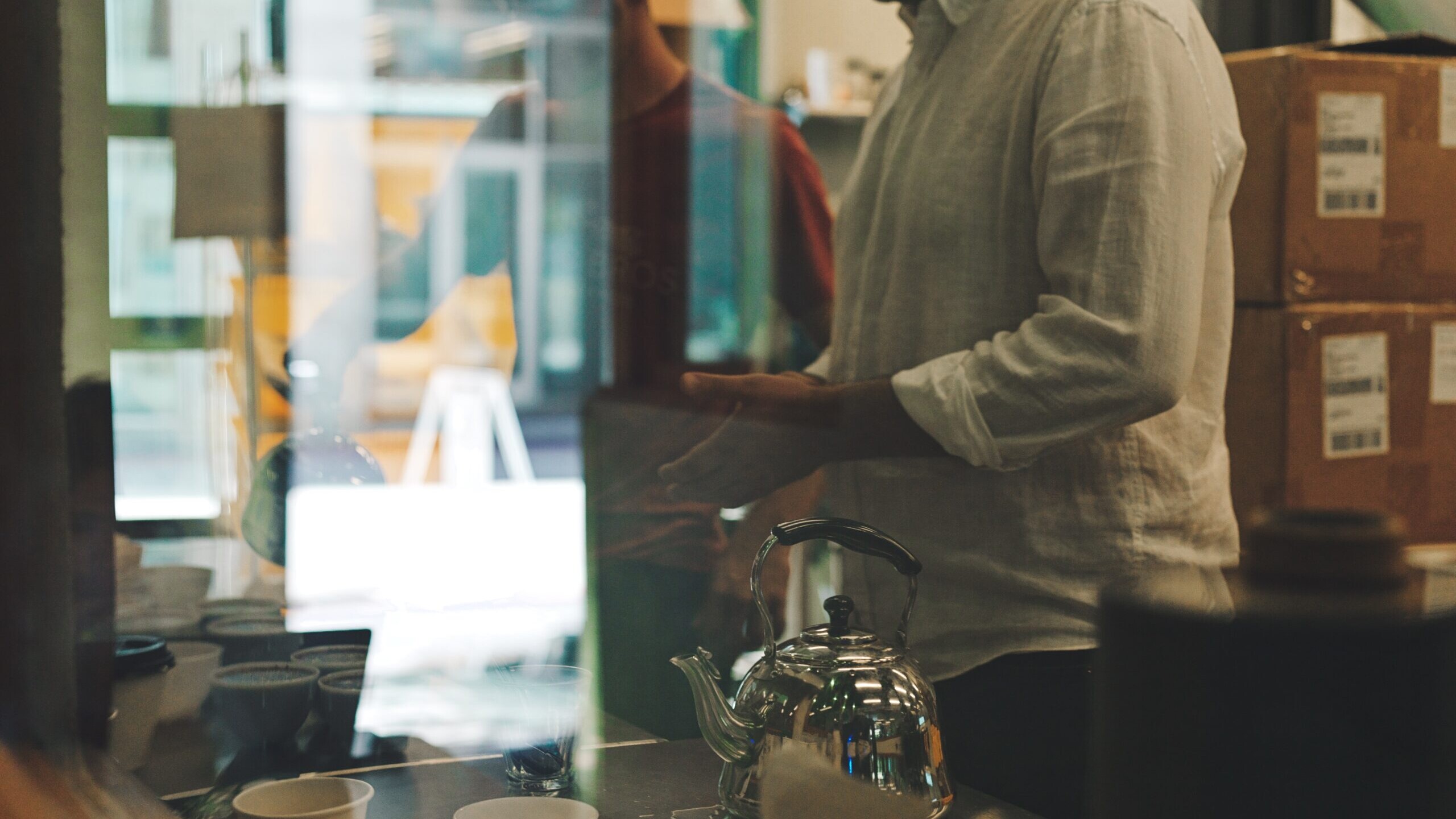 Two people prepare coffee behind a café counter with a metal kettle and tasting cups, viewed through a glass window with reflections.