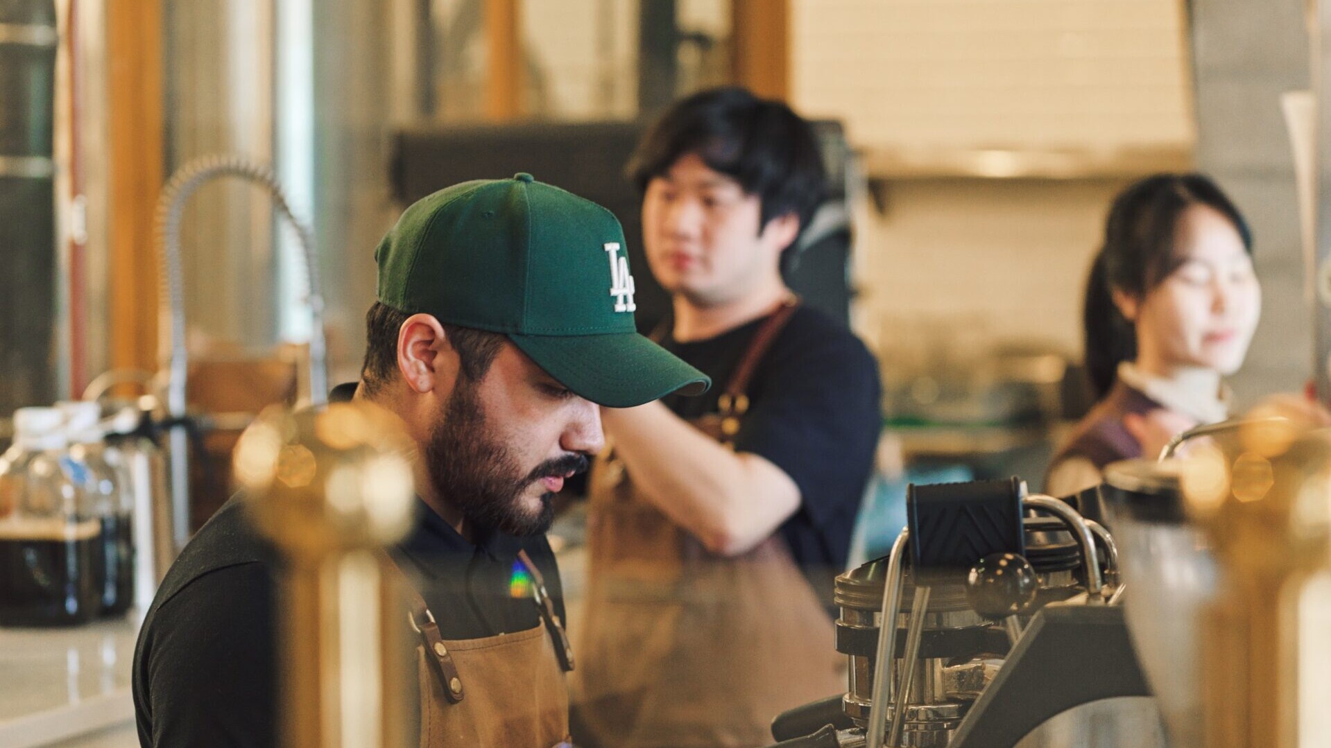 Barista in a green LA cap and brown apron works at an espresso machine in a busy café, with two coworkers blurred in the background.